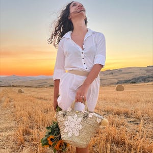 Puede incluir: Una mujer con un vestido blanco sostiene una bolsa de paja con un diseño de flor de crochet. La bolsa tiene un asa de cuero marrón y un pompón blanco. La mujer está de pie en un campo de balas de paja con una puesta de sol en el fondo.