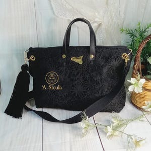 May include: Black tote bag with a gold embroidered design and the text "A Sicula". The bag has a black tassel and a long adjustable strap.