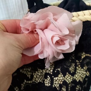 Pu&ograve; includere: Un fiore di tessuto rosa chiaro con petali a strati. Il fiore &egrave; posizionato sopra un tessuto di pizzo nero e un cestino intrecciato. I delicati petali del fiore sono di un rosa pastello tenue.