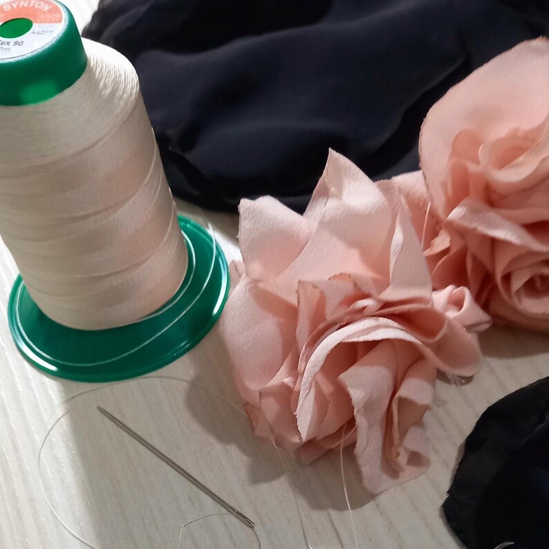 Pu&ograve; includere: Una bobina di filo rosa chiaro su una base verde, un ago e filo, e due fiori di tessuto rosa chiaro sono visibili. Il tessuto scuro sullo sfondo suggerisce un progetto di cucito o artigianato.