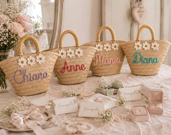 May include: Four woven straw tote bags with two handles each, decorated with three white daisy appliques and personalized names in cursive embroidery. The names read Chiara in purple, Anne in red, Mama in pink, and Diana in teal blue.