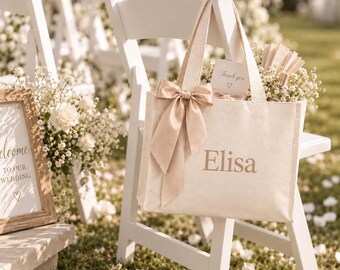 May include: A cream-colored canvas tote bag featuring the name Elisa printed in a metallic gold font. The bag is adorned with a large, tied champagne-colored satin ribbon bow on the front handle and contains a small card labeled Thank you.