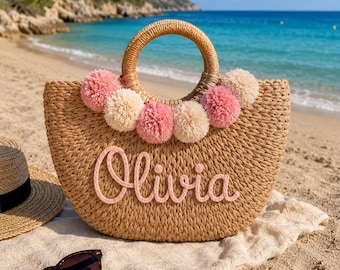 May include: A woven straw handbag with a semi-circular shape and two round handles. The bag features a row of alternating pink and cream-colored pom-poms along the top edge and the name Olivia written in pink script lettering on the front.