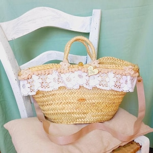 Puede incluir: Un bolso de mimbre con ribete de encaje blanco y pompones rosas. El bolso tiene una correa rosa y una etiqueta de madera con el nombre "Inge" en ella.