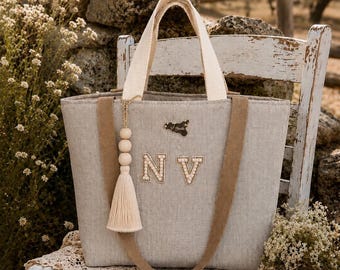 May include: A light gray tote bag featuring two beige fabric handles and a decorative tassel with wooden beads. The front displays the letters N and V in a pearl-embellished font and a small dark metal emblem shaped like the island of Sicily.