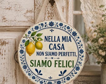 May include: A round decorative sign with a cream background, blue floral border, and two yellow lemons on a branch. The text reads: NELLA MIA CASA NON SIAMO PERFETTI, SIAMO FELICI. The sign hangs from a beige ribbon attached to a metal handle on each side.