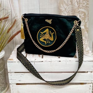 Puede incluir: Un bolso de mano de terciopelo verde oscuro con una correa de cadena dorada y una correa para el hombro estampada. El bolso presenta un emblema bordado en oro y una borla dorada. Un mapa dorado de Sicilia está en la esquina superior derecha del bolso.