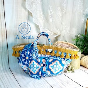 Puede incluir: Una bolsa de mano azul y blanca con estampado, con borlas amarillas y un asa de paja. La bolsa tiene una bufanda azul y blanca con estampado colgando de ella. La bolsa está etiquetada como "A' Sicula".