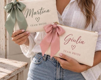 May include: Two off-white canvas zipper pouches featuring a gold zipper and a decorative satin bow. One pouch has a sage green bow and the text Martina and TEAM BRIDE in green. The other has a dusty rose bow and the text Giulia and TEAM BRIDE in rose.