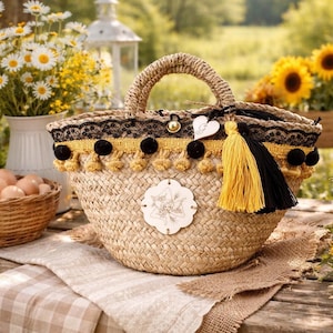 Puede incluir: Un bolso tote de paja tejida con forma redondeada, con asa trenzada y adornos decorativos. El adorno incluye encaje negro, borlas amarillas y pompones negros. Un adorno de madera con un diseño floral está adherido al frente. Una borla amarilla y negra cuelga de un lado.