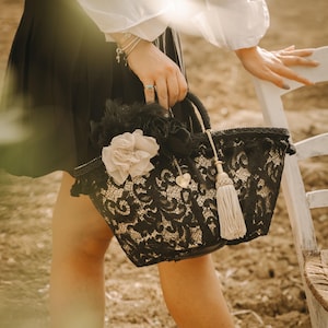 May include: A black lace tote bag with a white tassel and a large white flower embellishment. The bag has a heart-shaped charm.