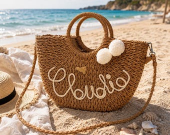 May include: A brown woven straw handbag with a semi-circular shape, featuring the name Claudia embroidered in white script on the front. The bag includes two round top handles, a long shoulder strap, and two white pom-poms attached to the side.