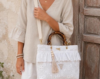 May include: A white eyelet lace handbag featuring a bamboo handle, a detachable beige canvas shoulder strap, and a ruffled trim. The bag is adorned with a beige tassel, wooden beads, and a letter V patch, with gold-tone hardware accents.