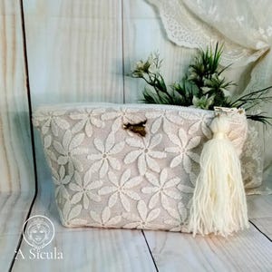 Può includere: Una pochette bianca con cerniera, con un designo ricamato floreale e un fiocco bianco. La pochette è su una superficie di legno.