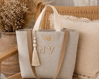 May include: A light gray linen tote bag featuring two beige fabric handles and a decorative tassel with wooden beads. The front of the bag displays the letters NV in white pearl-like embellishments and a small gold-toned map-shaped charm.