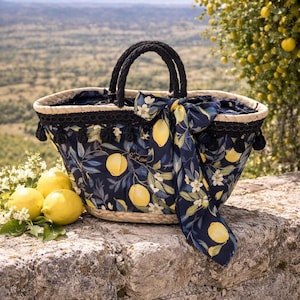 May include: A woven straw tote bag with a navy blue fabric lining and a lemon print. The bag has black rope handles and a matching scarf tied around it. Several lemons and white flowers are next to the bag.