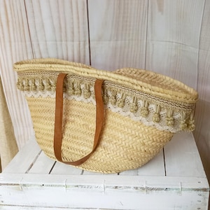 Puede incluir: Un gran bolso de paja tejido con asas de cuero marrón y un ribete decorativo de encaje beige y borlas.