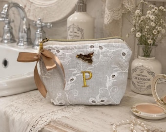 May include: A light gray fabric pouch featuring white floral embroidery, a gold-colored letter P, and a small gold-colored map-shaped charm. A tan ribbon is tied to the gold zipper pull. The pouch has a structured shape and a top zipper closure.