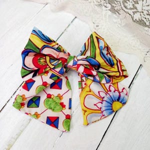 May include: A colourful bow made of fabric with a geometric and floral pattern. The bow is tied in a knot and has a white background with red, blue, yellow, and green accents.