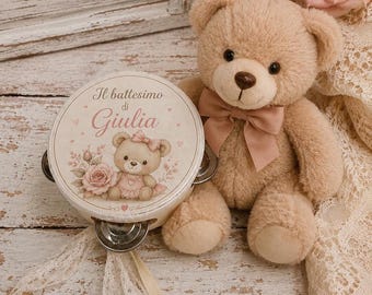 May include: A beige plush teddy bear with a dusty pink ribbon bow around its neck, positioned next to a round tambourine. The tambourine features a printed illustration of a teddy bear, a pink rose, and the text Il battesimo di Giulia.