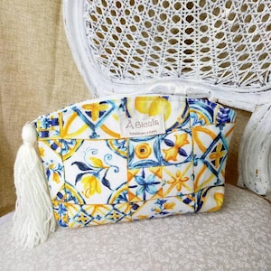 Può includere: Una pochette bianca con cerniera e un pon pon. La pochette è decorata con un motivo colorato di piastrelle blu, gialle e bianche. La pochette ha un'etichetta che recita "A Sicilia" con il testo "Realizzato a Mano" sotto.