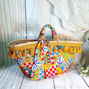 Può includere: Una grande borsa a cestello in vimini gialla con una fodera in tessuto a fantasia colorata e un bordo con frange gialle. La borsa ha un'unica maniglia rivestita in tessuto e un ciondolo a forma di cuore.