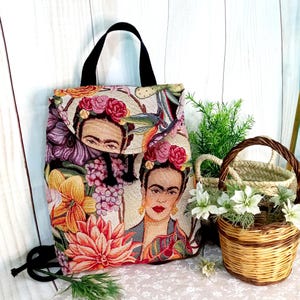 Può includere: Uno zaino piccolo e floreale con una tracolla nera e una chiusura a lembo. Lo zaino presenta un design a tappezzeria colorato con immagini di Frida Kahlo e fiori.