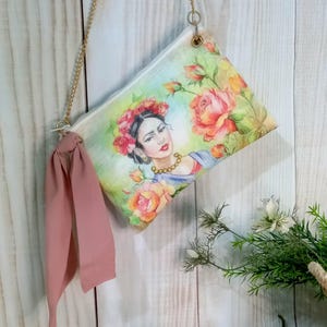 Può includere: Una borsa di tela bianca con un design floreale e un ritratto di una donna con una corona di fiori. La borsa ha una tracolla a catena dorata e un nastro rosa.