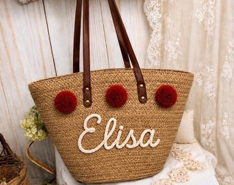 May include: A woven straw tote bag featuring two brown leather shoulder straps and three red pom-poms attached to the front. The name Elisa is written in white cursive script lettering across the center of the bag, which has a wide base.