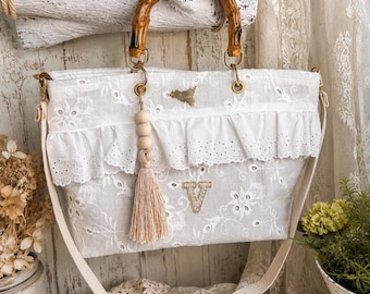 May include: A white tote bag featuring an intricate floral eyelet lace pattern, a ruffled trim, and a letter V patch. The bag includes a bamboo handle, a long shoulder strap, and a hanging tassel ornament with wooden beads and a gold charm.