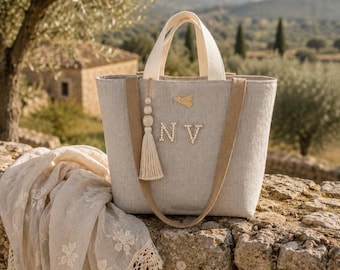 May include: A light gray linen tote bag featuring two cream-colored top handles and two tan shoulder straps. The front is decorated with the letters NV in white, a small gold-toned emblem, and a long cream-colored tassel hanging from the handle.