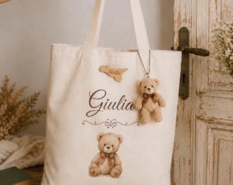 May include: A cream-colored canvas tote bag featuring the name Giulia in brown script, a printed illustration of a seated teddy bear, a wooden cutout shaped like Sicily, and a small plush teddy bear keychain attached to the strap.