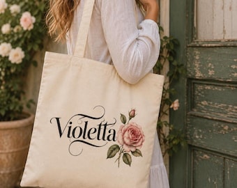 May include: A cream-colored canvas tote bag featuring the name Violetta written in a black, elegant, cursive script font. To the right of the text is a detailed illustration of a single pink rose with green leaves and a stem.
