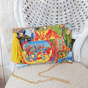 Può includere: Una pochette colorata con una tracolla a catena dorata e un fiocco giallo. La borsa presenta un design a patchwork con una scena di persone in abiti medievali.