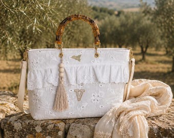 May include: A white eyelet lace handbag featuring a bamboo handle, a ruffled trim, and a beige tassel with wooden beads. The bag displays a small gold-toned map of Sicily and a letter V embroidered in a light beige thread on the front panel.