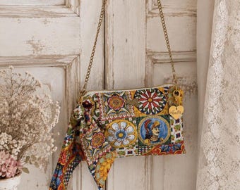 May include: A rectangular crossbody bag featuring a vibrant, multi-colored tile-inspired pattern with floral motifs and a knight illustration. It includes a gold-tone chain strap, a matching patterned fabric tie, and a heart-shaped gold charm.
