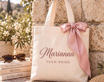 May include: A cream-colored canvas tote bag featuring the name Marianna and the text TEAM BRIDE in a dark pink script and serif font. The bag is adorned with a large, dusty pink satin ribbon tied in a bow on the upper right handle attachment.