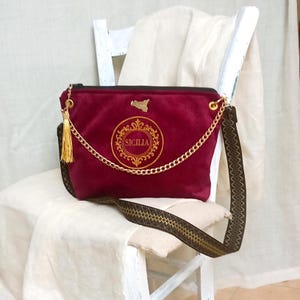 May include: A burgundy velvet shoulder bag with a gold chain strap and tassel. The bag features gold embroidery with the word "SICILIA" and a gold outline of Sicily. The bag is on a white chair.