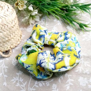 Op de afbeelding: Een gele, blauwe en witte geprinte scrunchie haarband. De scrunchie is gemaakt van stof en heeft een gerimpeld ontwerp.