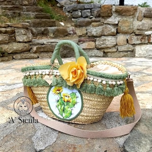 May include: A straw tote bag with a green and white trim, a yellow flower, and a painted cactus design. The bag has a long strap and a small wooden tag that says "Chiana".