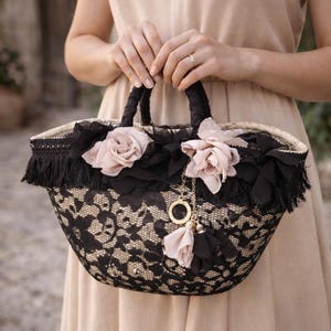 Pu&ograve; includere: Una borsa tote in pizzo nero e beige con frange e manici neri. La borsa &egrave; decorata con rose di tessuto rosa e un ciondolo dorato con nappa. La borsa &egrave; tenuta da una persona.