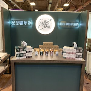 May include: A trade show booth featuring a dark green backdrop with a circular illuminated sign reading Super Fat, and a rectangular sign reading Feel Good Fat. The booth displays stacks of nut butter boxes and small pouches on a wooden counter.