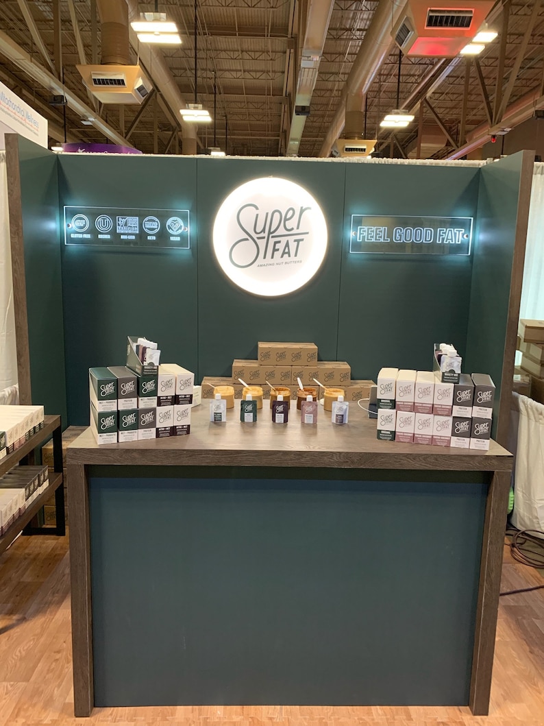May include: A trade show booth featuring a dark green backdrop with a circular illuminated sign reading Super Fat, and a rectangular sign reading Feel Good Fat. The booth displays stacks of nut butter boxes and small pouches on a wooden counter.