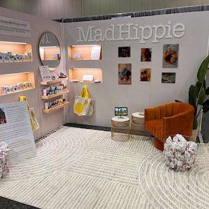 May include: A trade show booth for MadHippie featuring light-colored walls with recessed shelving displaying skincare products, a round mirror, a burnt orange velvet armchair, two wire side tables, a large area rug, and two wire baskets filled with items.