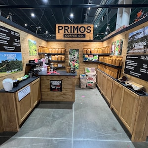 May include: A Primos Coffee Co. booth featuring light wood paneling, shelves stocked with brown coffee bags, and black menu boards. The central counter displays the text Primos Coffee Co. We're sellin' what you're smellin' and a large burlap sack.