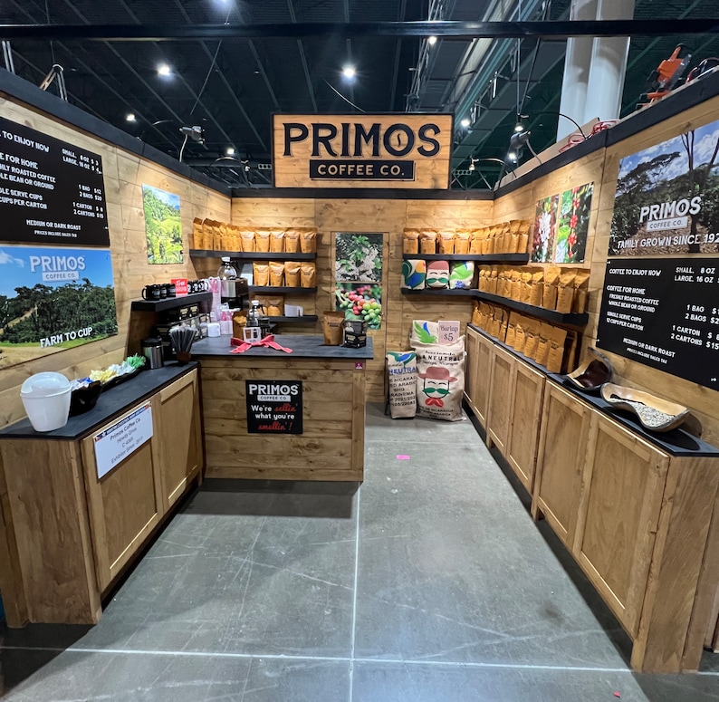 May include: A Primos Coffee Co. booth featuring light wood paneling, shelves stocked with brown coffee bags, and black menu boards. The central counter displays the text Primos Coffee Co. We're sellin' what you're smellin' and a large burlap sack.