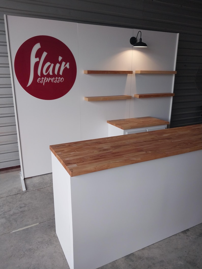 May include: A white display counter with a light wood top and a matching white backdrop featuring a red circular logo with the text flair espresso. The setup includes four light wood floating shelves and a black wall-mounted lamp fixture.