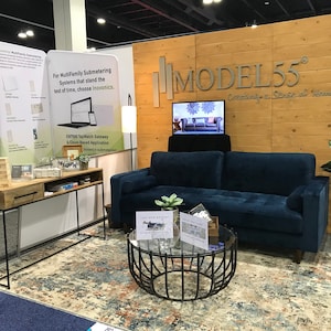 May include: A navy blue tufted sofa with a round glass coffee table featuring a black metal base, a wooden console table with a black metal frame, and a patterned area rug. The display includes a wooden wall sign reading MODEL55 Creating a Sense of Home.