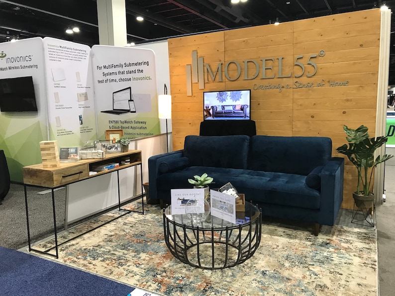 May include: A navy blue tufted sofa with a round glass coffee table featuring a black metal base, a wooden console table with a black metal frame, and a patterned area rug. The display includes a wooden wall sign reading MODEL55 Creating a Sense of Home.