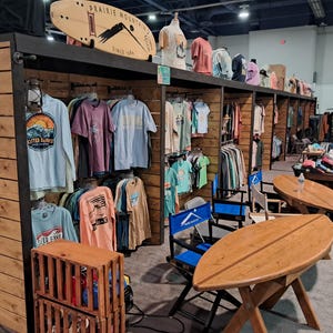 May include: A retail display booth featuring wooden shelving units filled with various graphic t-shirts, a surfboard-shaped sign labeled Prairie Mountain, and two surfboard-shaped wooden tables with blue director chairs in a large indoor venue.